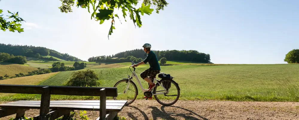 Ein Radfahrer auf einem E-Bike fährt einen ländlichen Weg entlang, umgeben von grünen Hügeln und Feldern an einem sonnigen Tag. Im Vordergrund ist eine hölzerne Bank zu sehen, während die Landschaft im Hintergrund Weite und Ruhe vermittelt.