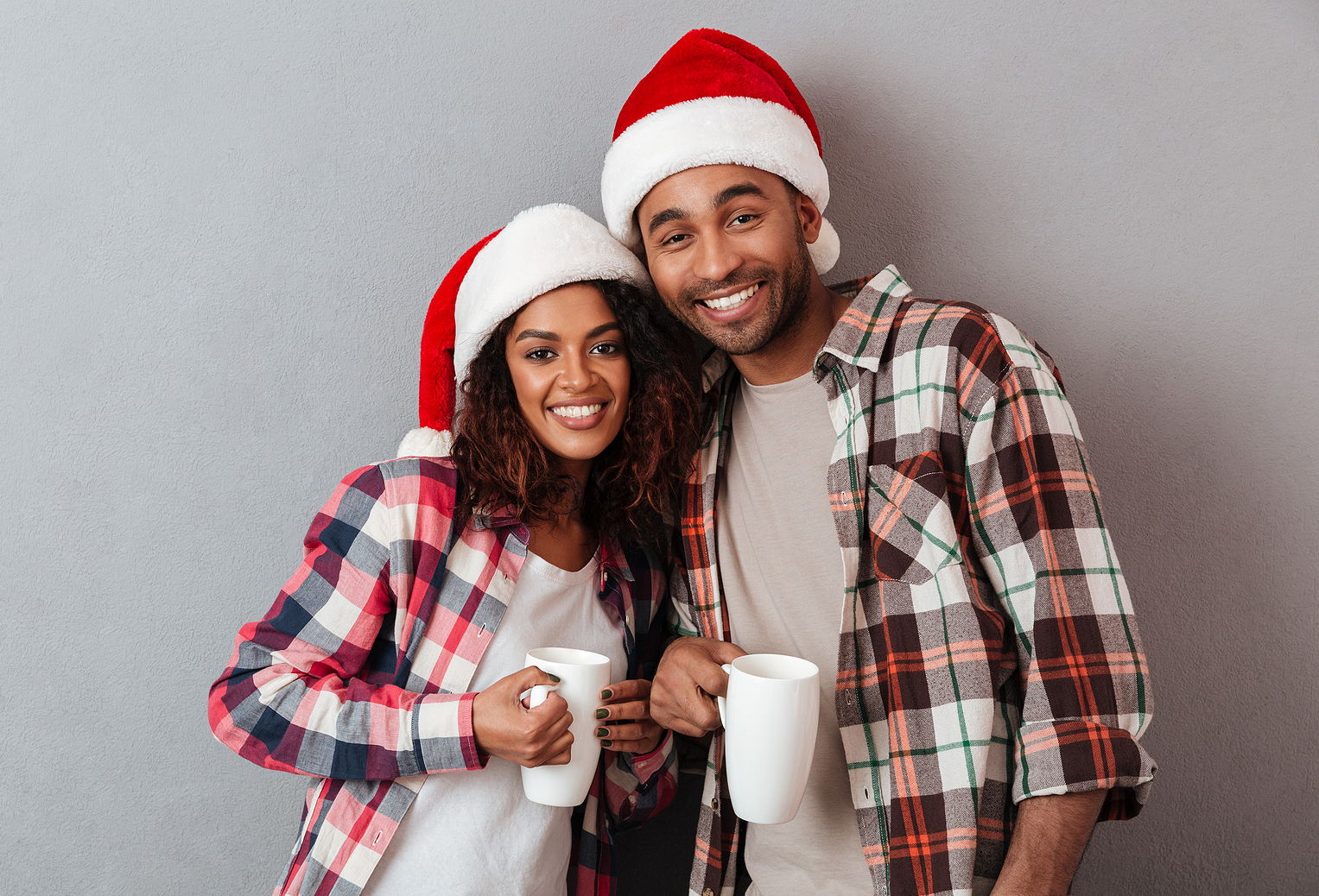 An african american man and woman smile together wearing santa hats and holding a mug.