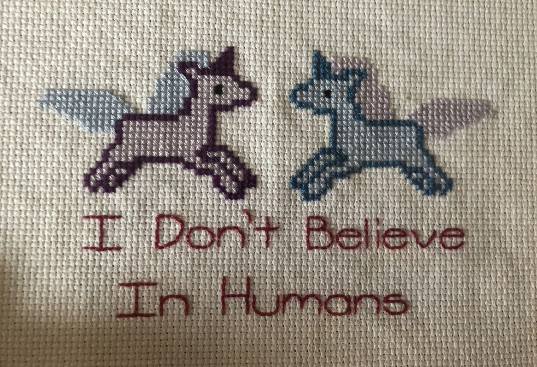 Stitching of two unicorns facing eachother with the words I don't believe in humans, underneath them.