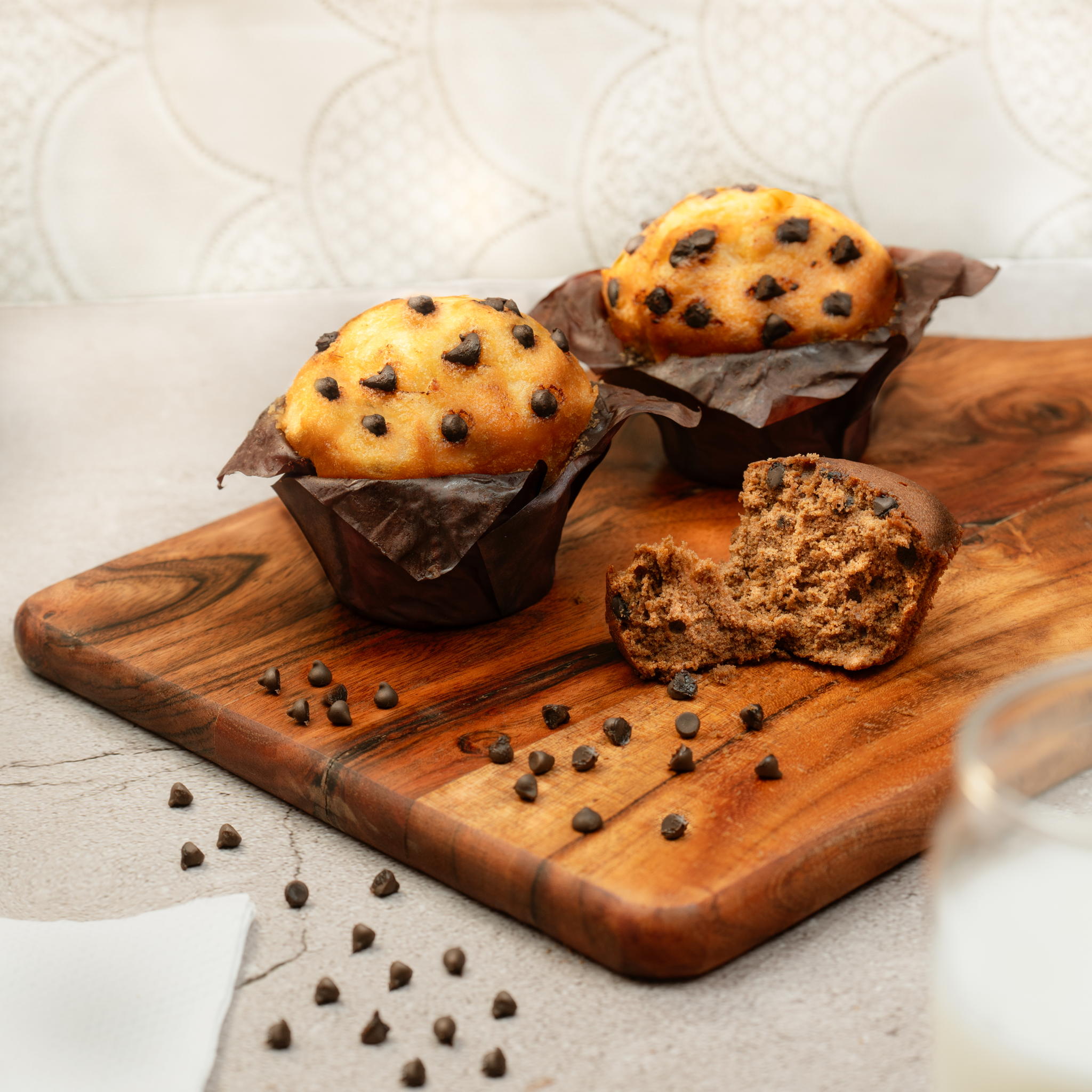 Two chocolate chip muffins, one with a bite taken out, on a wooden cutting board