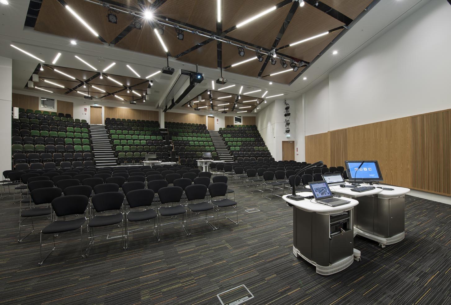Modern lecture hall with tiered seating, mostly black chairs, green accents, and two podiums with computers and microphones at the front. The room features wooden panels and geometric ceiling lights.