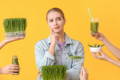 A woman in front of a yellow background surrounded by different forms of wheat grass: smoothie, juice, fresh grass sprigs, etc. 