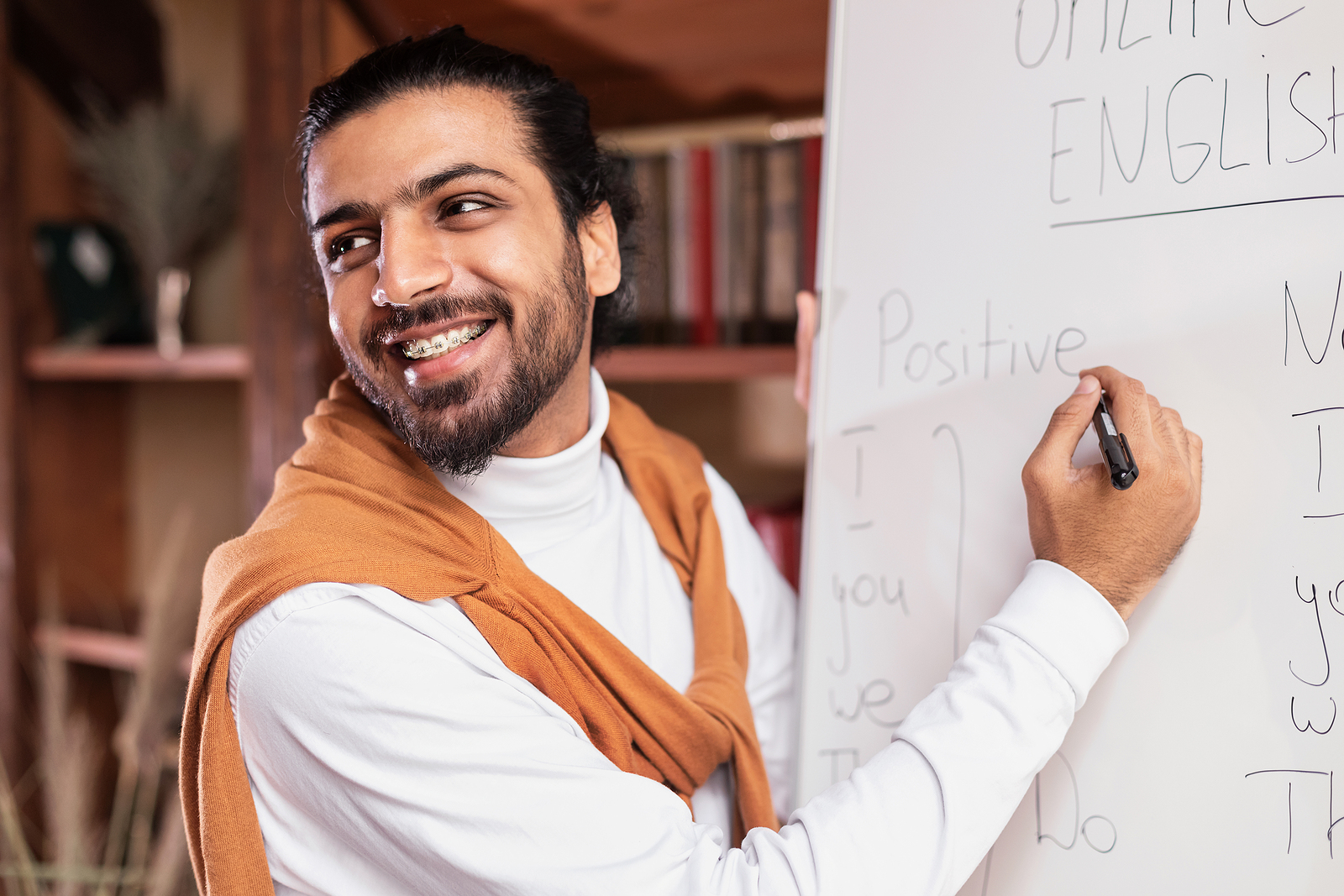 A multi racial man with braces and his hair in a bun, smiling and looking to the audience, writing words on a white board.