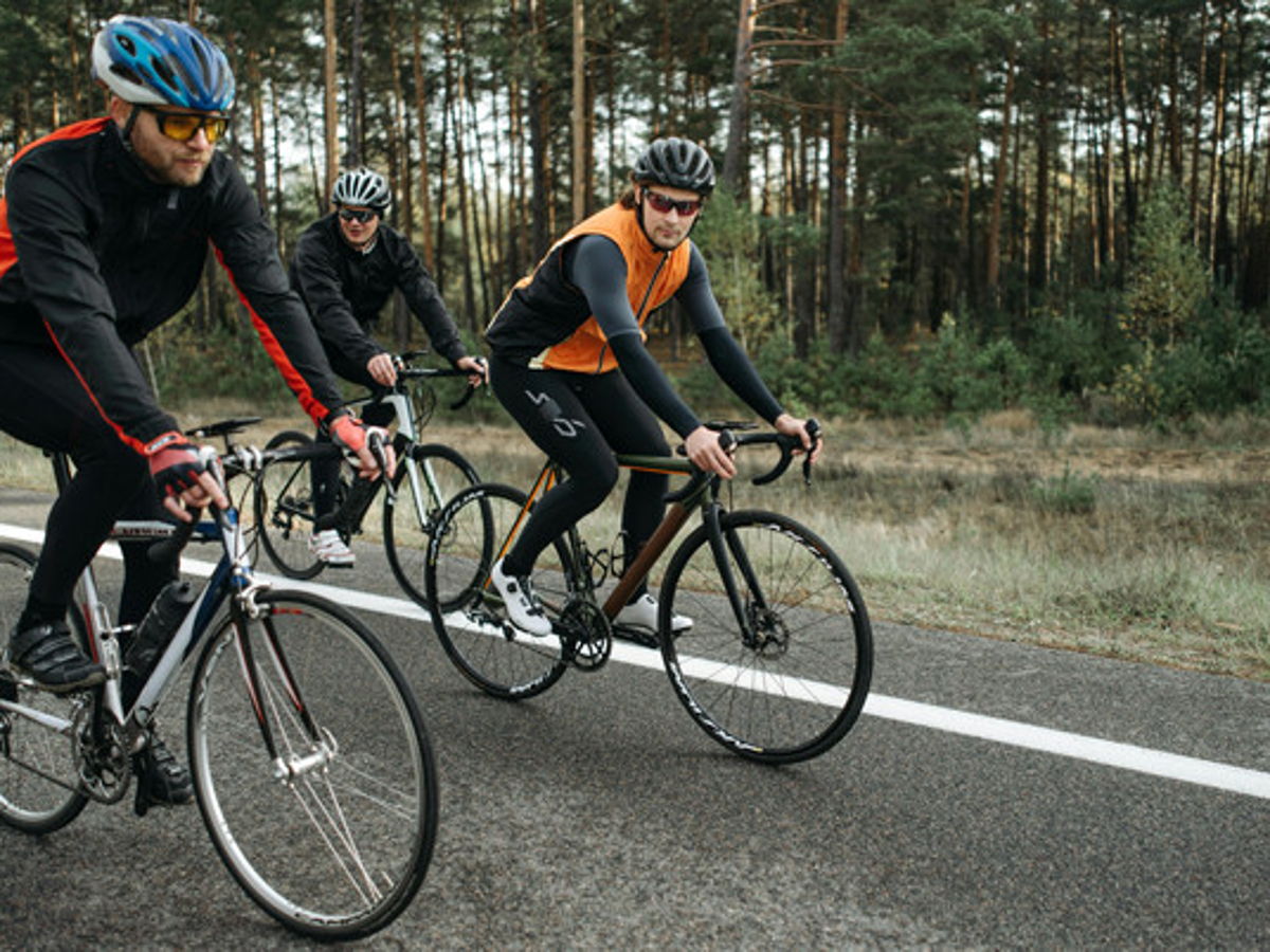Drei Radfahrer mit Rennrädern auf Landstraße im Wald mit Nadelbäumen im Hintergrund 