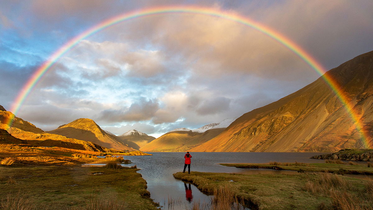 Person standing at the edge of Wast Water, watching a rainbow in cloudy sky.