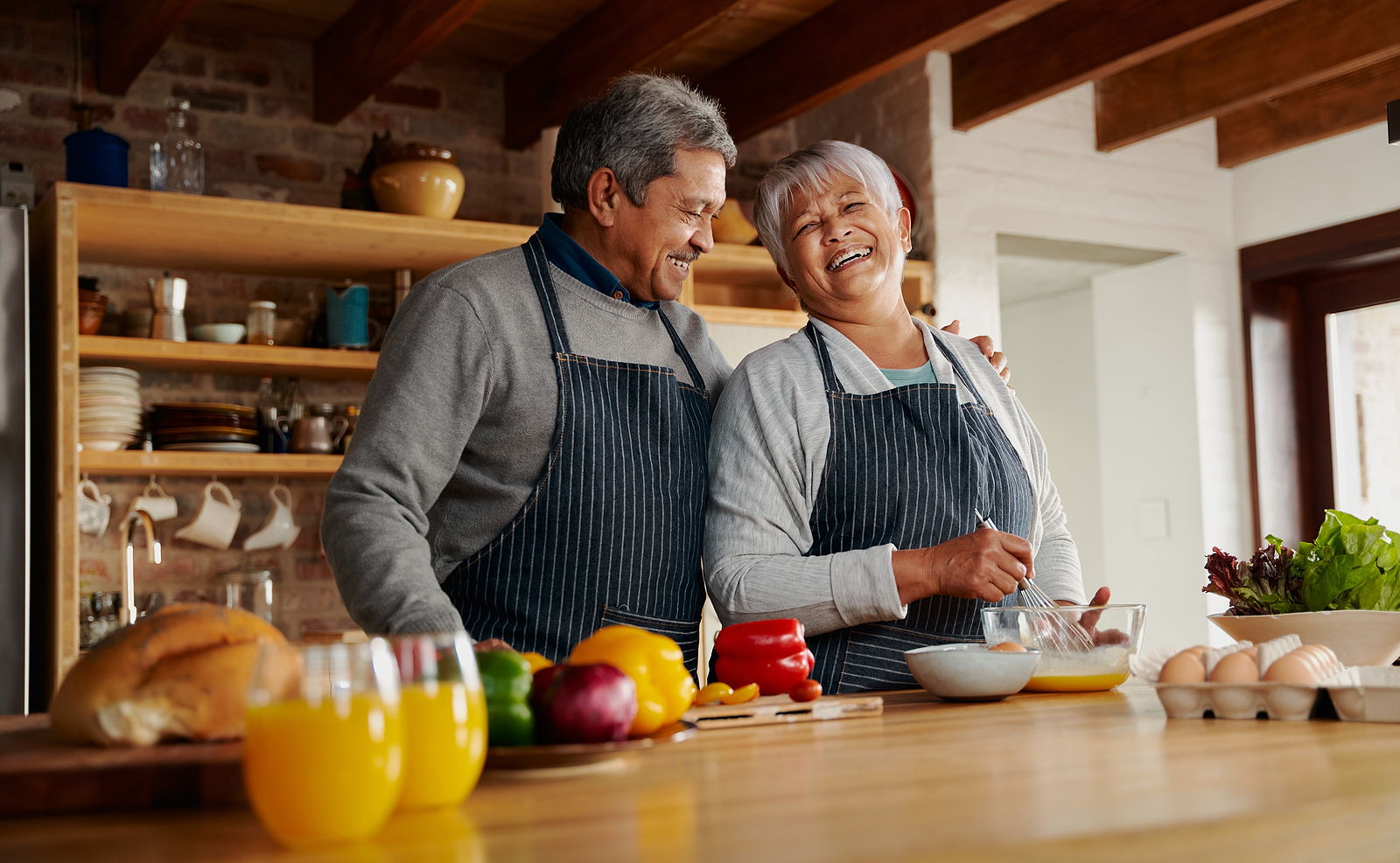 A bi racial married couple laugh together while making breakfast int heir kitchen. The husband has his hand on his wife's shoulder.