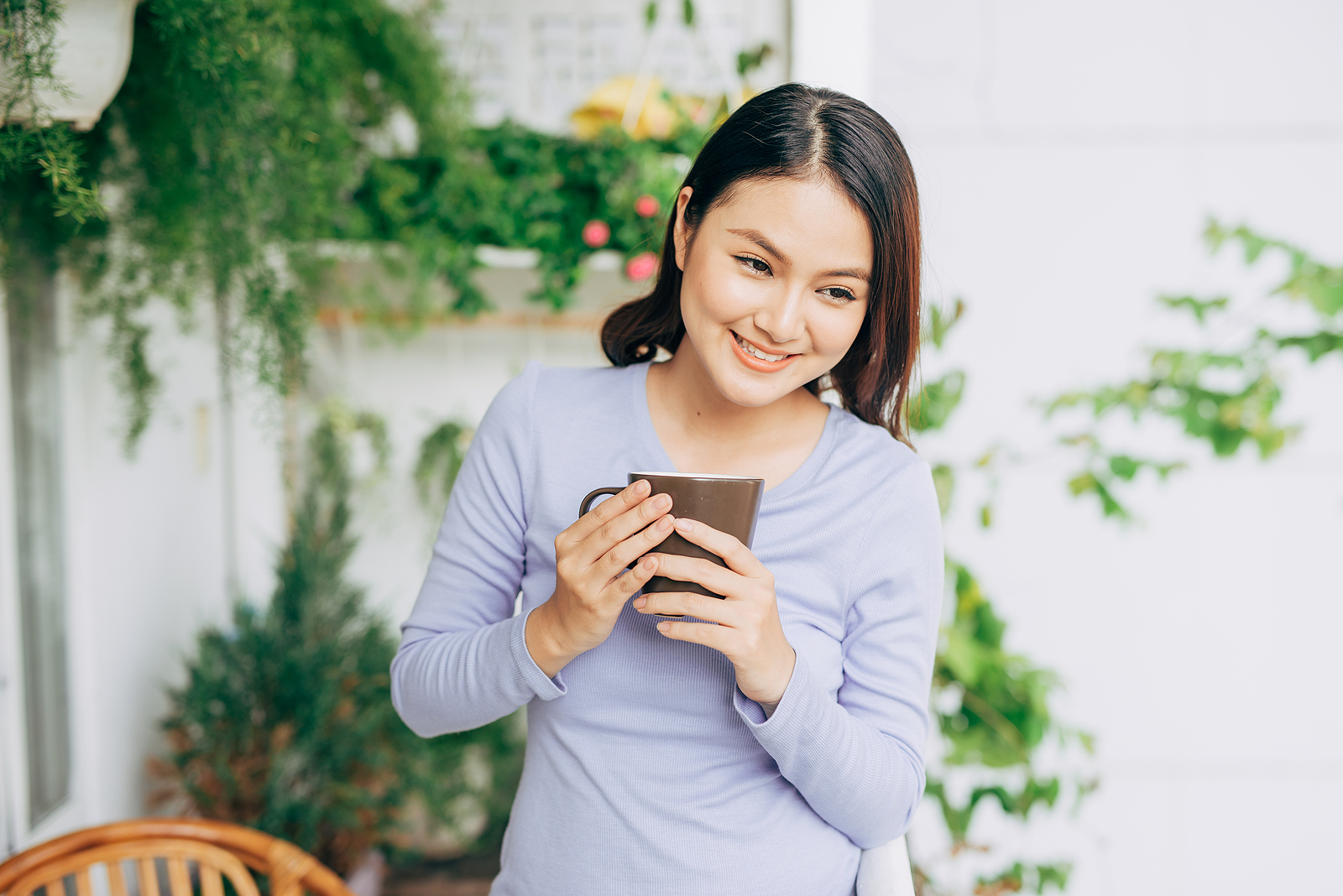 Image of an asian woman wearing a purple longsleeve shirt smiling and holding her coffee mug with both hands looking to the side. Behind her there are many plants on a white background.