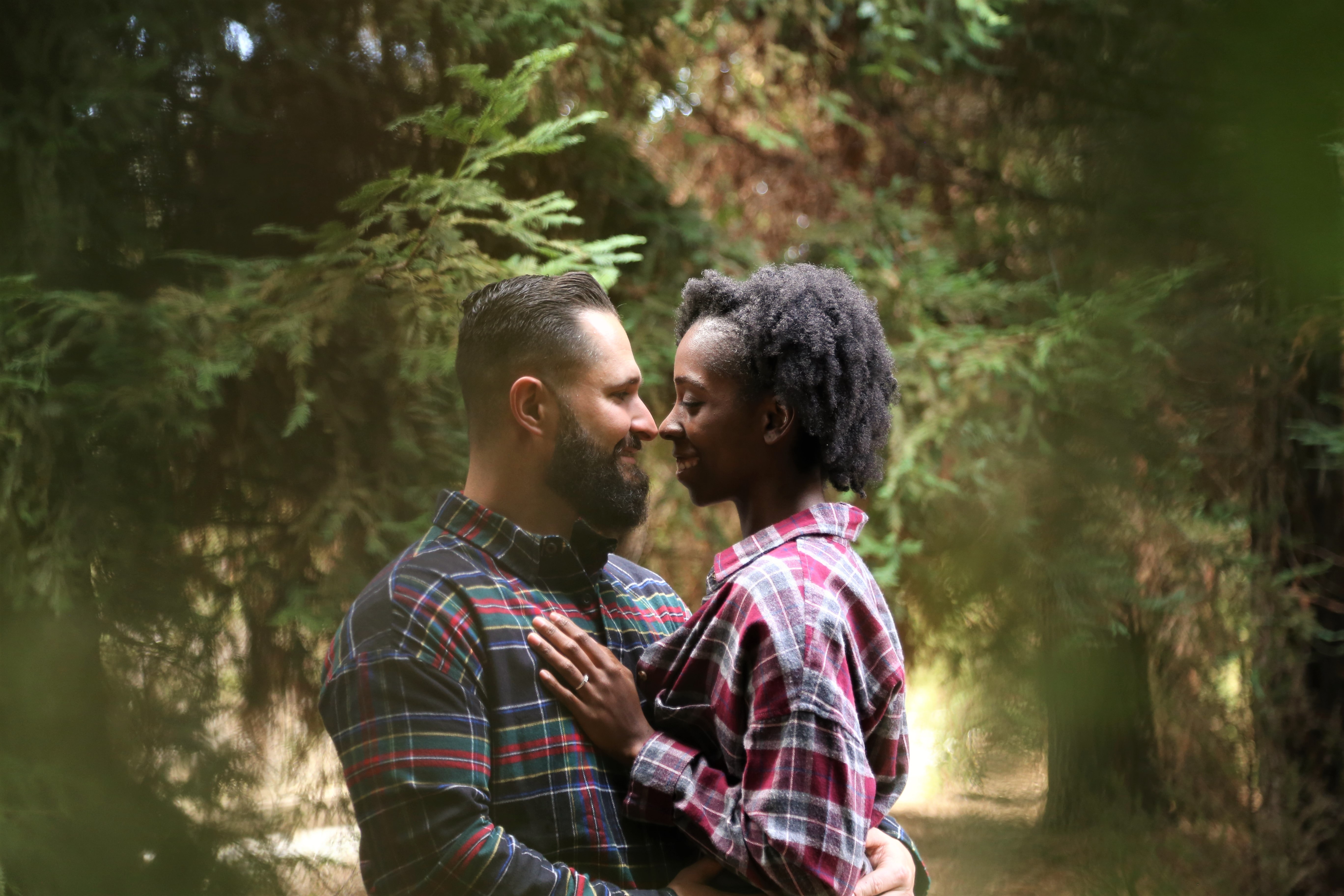 A multi racial couple shares an intimate moment near the woods, holding eachother and smiling.