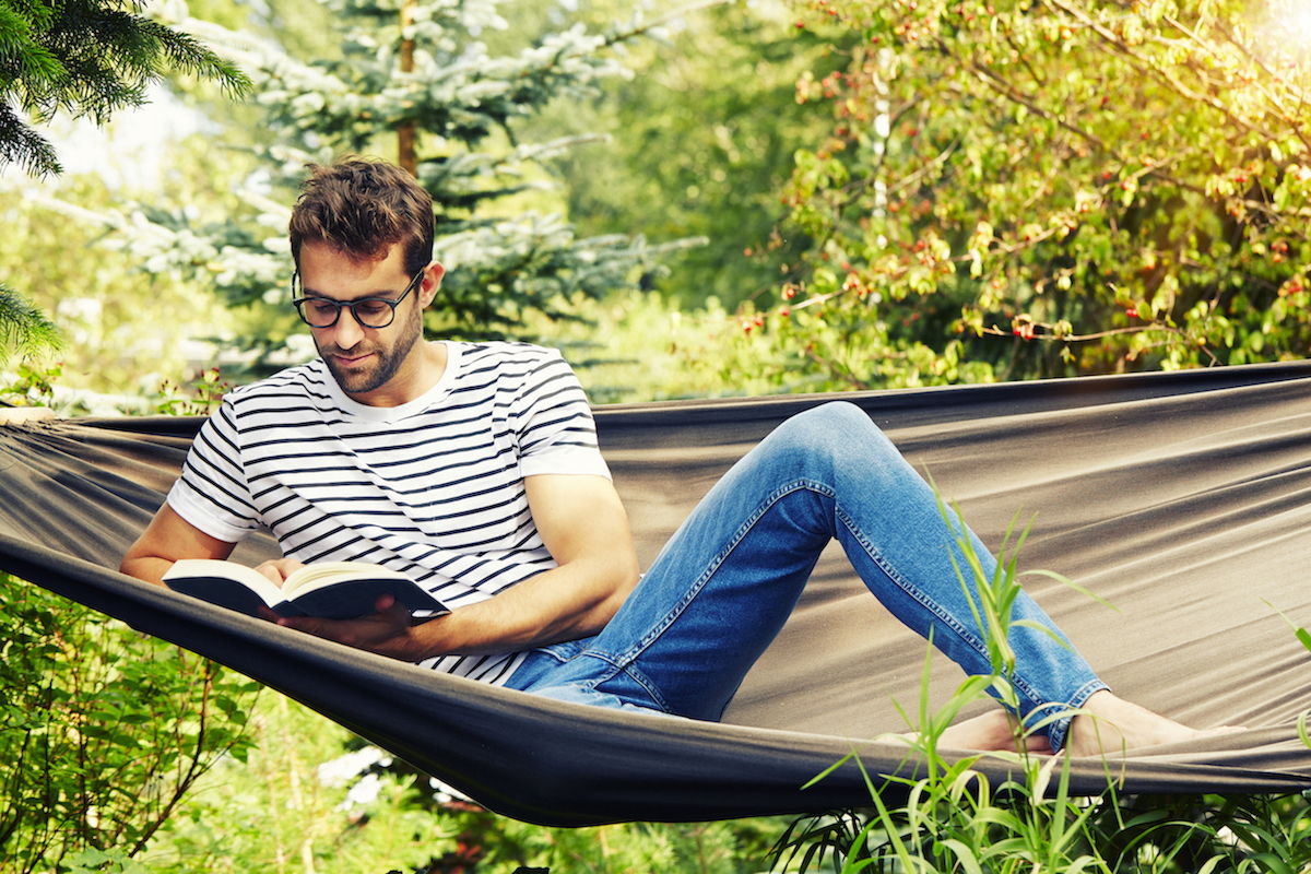 A hip young man with glasses lays on a hammock while reading a book.