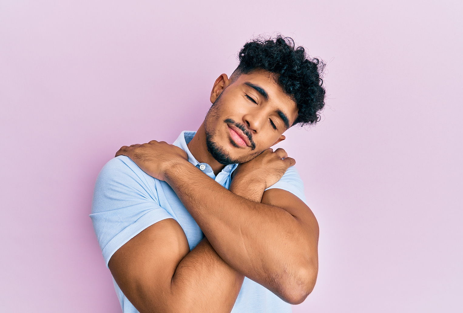 A multi ethnic man wearing a polo shirt hugs himself while smiling and eyes closed.