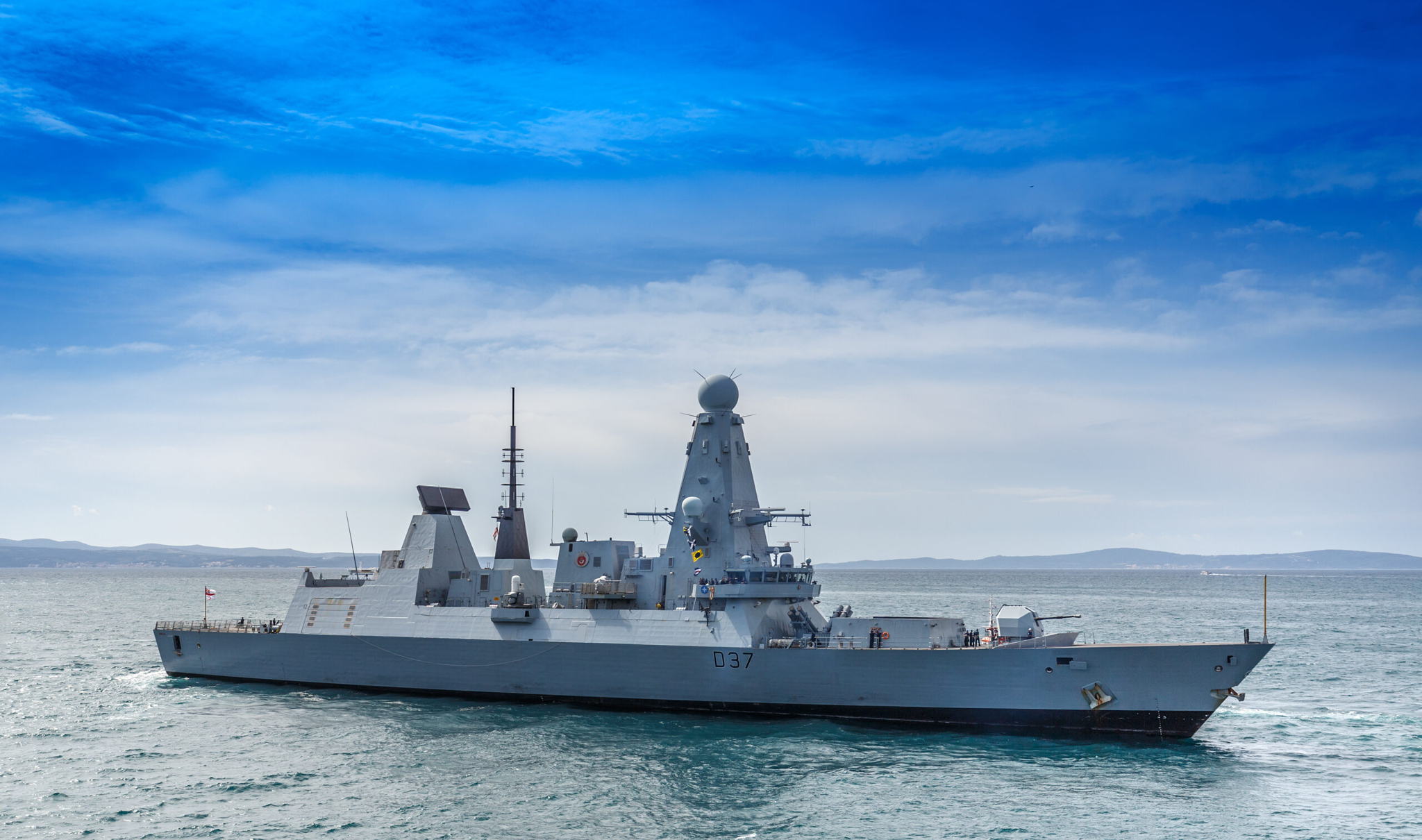 A modern naval warship sails on calm sea waters under a blue sky with scattered clouds; land is visible in the distant background.
