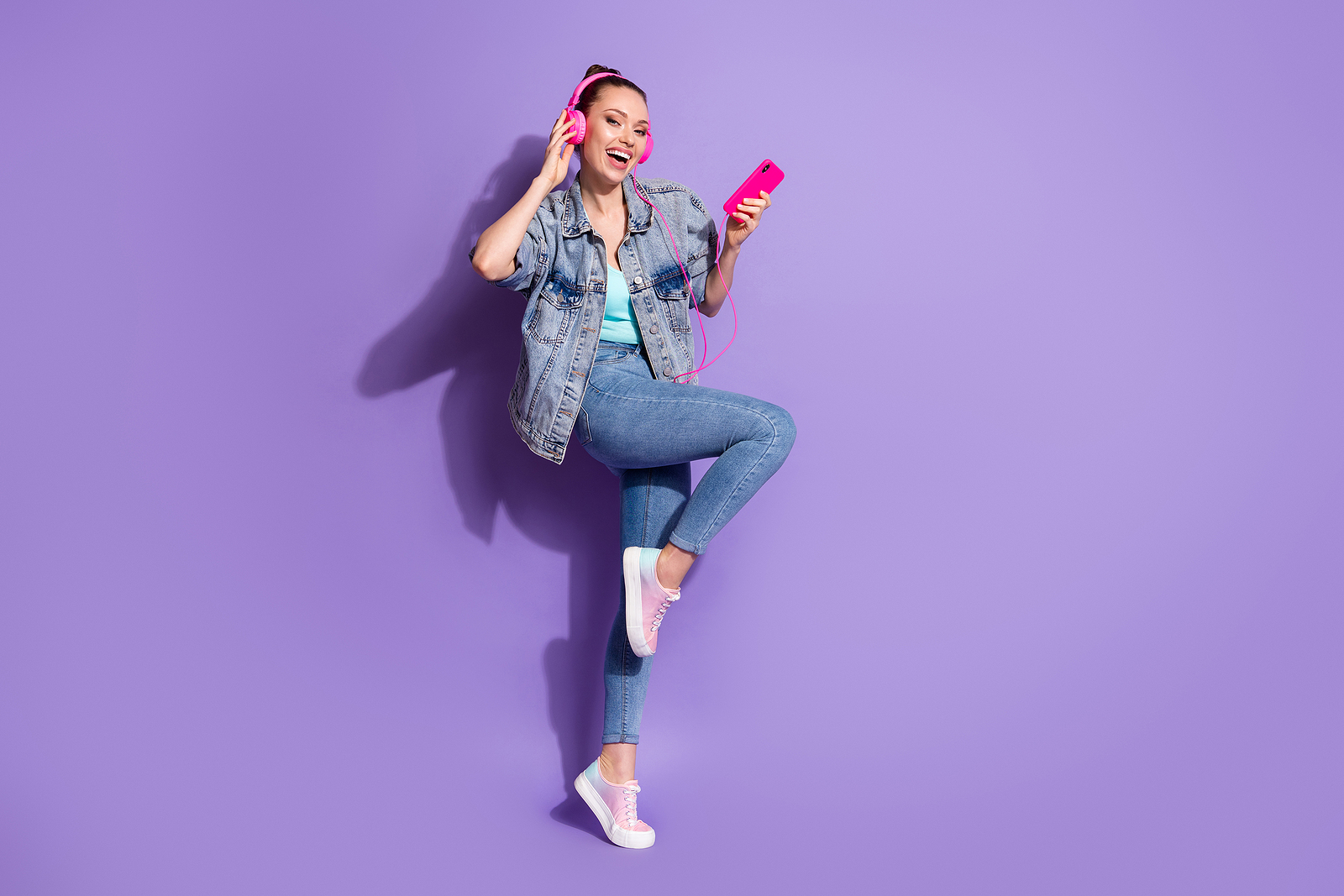 An attractive woman wearing pink headphones against a purple background, jumping in the air and smiling.