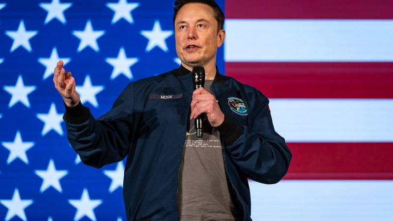 Elon Musk standing in front of an American flag background and speaking into a microphone.