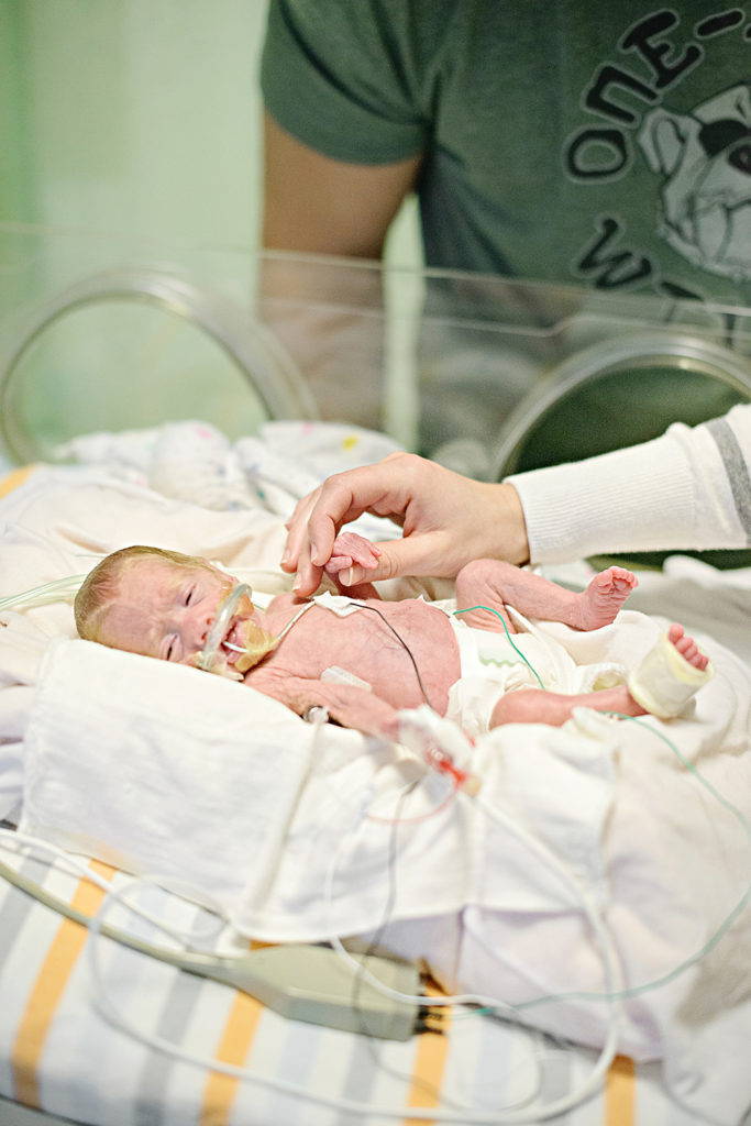 preemie baby with oxygen and OG tube in incubator in NICU holding mother's hand