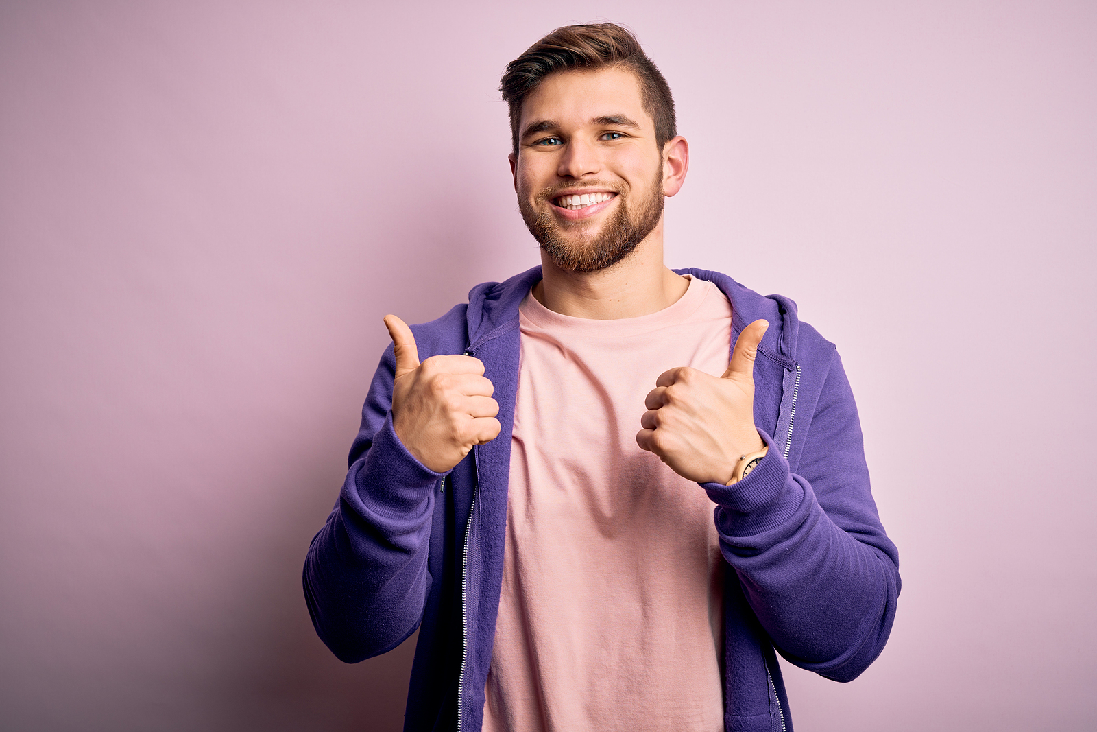 A young attractive man smiles showing thumbs up with both hands against a plain background.