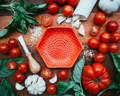 A red garlic grate plate sits among a colorful spread of basil, tomatoes, salt, garlic and a wooden tool