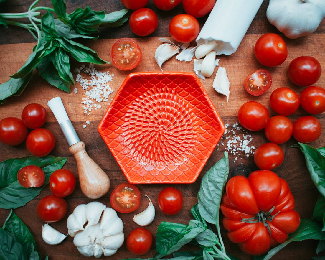A red garlic grate plate sits among a colorful spread of basil, tomatoes, salt, garlic and a wooden tool