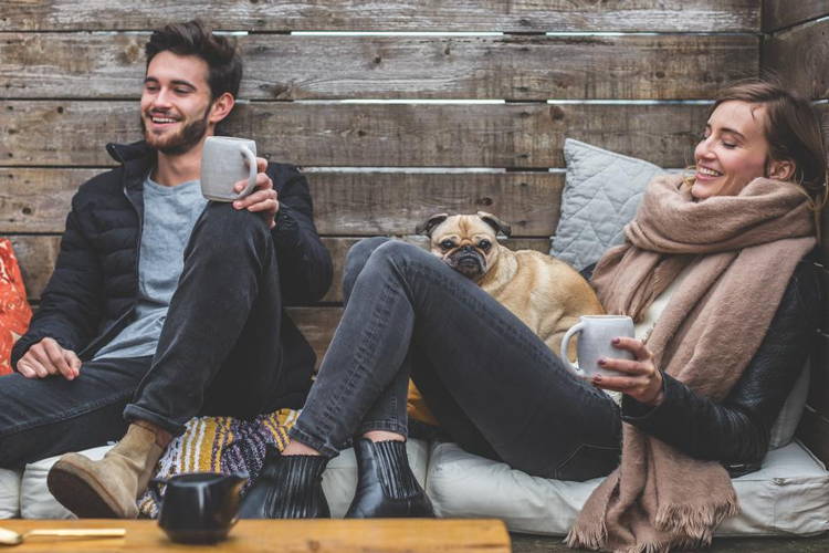 man and woman smiling with coffee mugs and pug on a bench