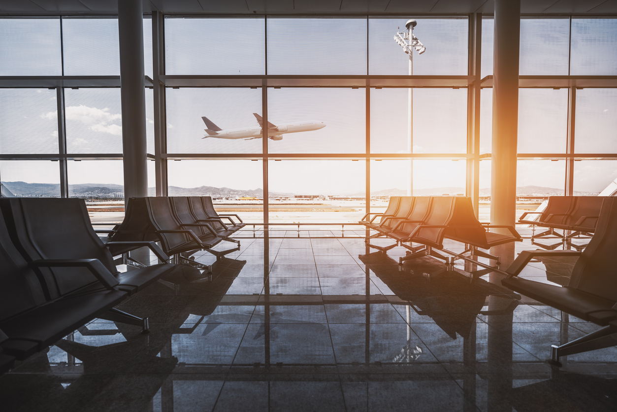 An airplane takes off outside large glass windows of an empty airport terminal, with rows of black seats and sunlight streaming in, reflecting on the shiny floor.