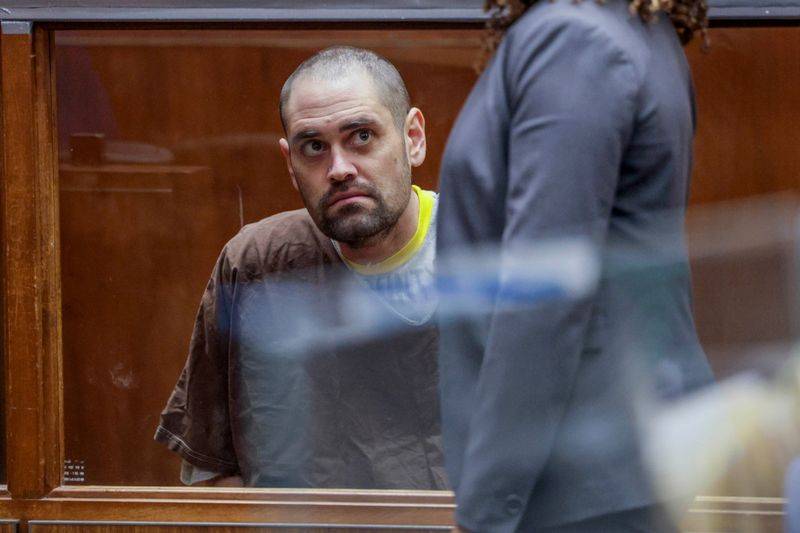 Nick Reiner sits behind a glass barrier in a courtroom. He is wearing a brown prison uniform on top of a yellow T-shirt visible at the collar. His head is shaved and his beard is closely trimmed. His mouth is turned down and his eyes are wide in an attent