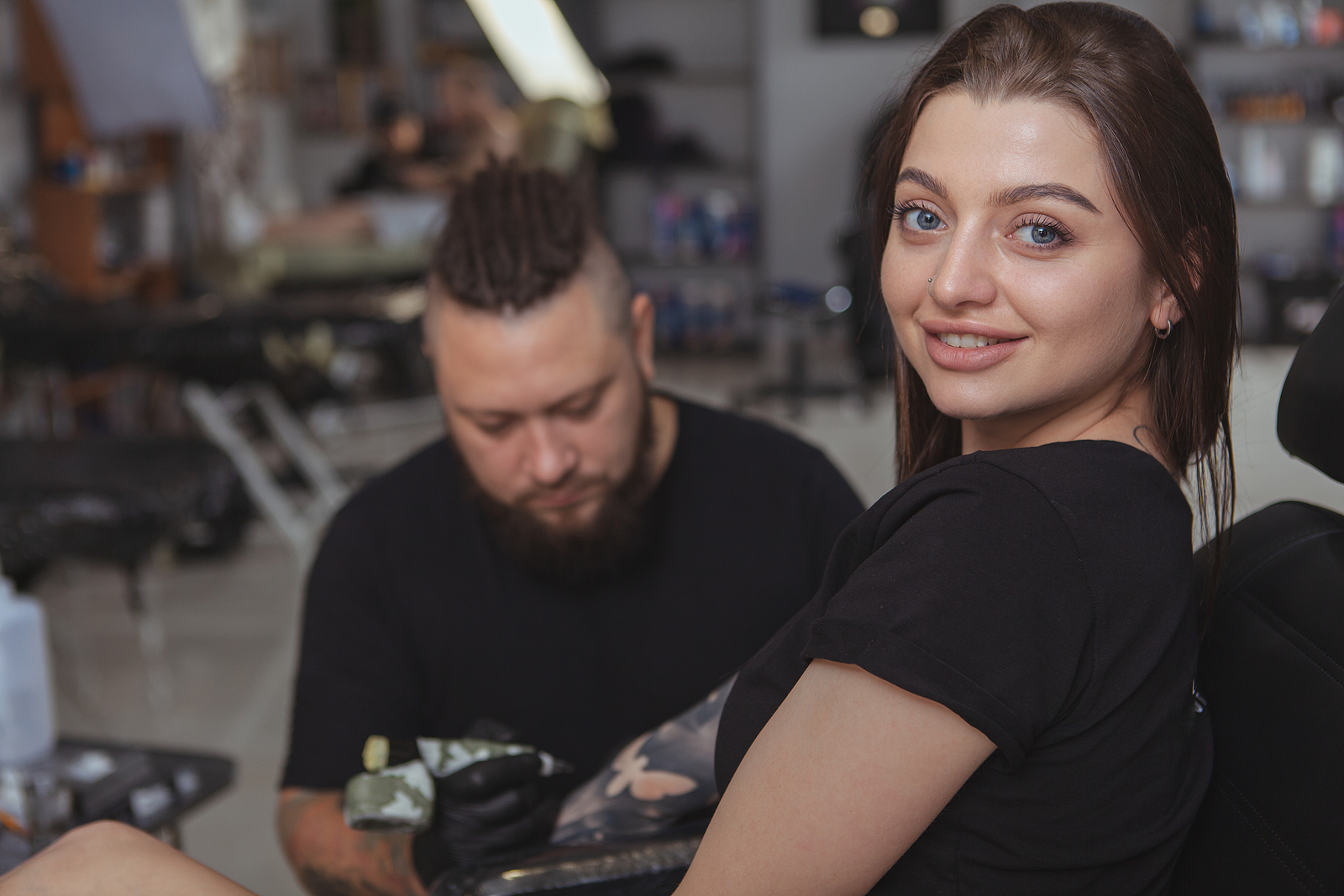A woman smiles and looks at the camera while getting a tattoo on her arm.