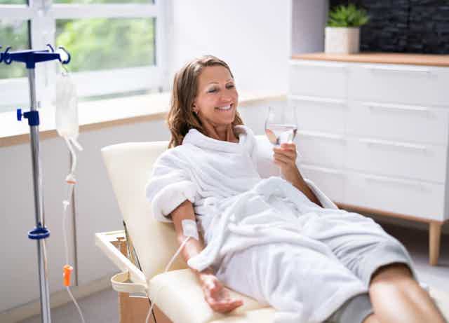 Woman in a white robe reclines in a spa chair, smiling and holding a champagne glass while receiving IV therapy.