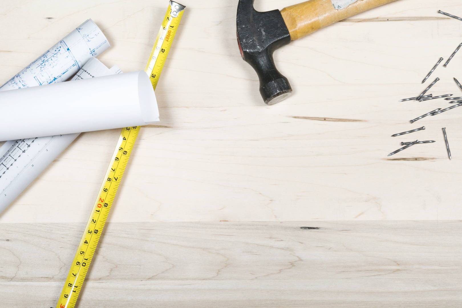 Planuri de construcție rulate, o ruletă, un ciocan și câteva cuie sunt dispuse pe o suprafață din lemn.