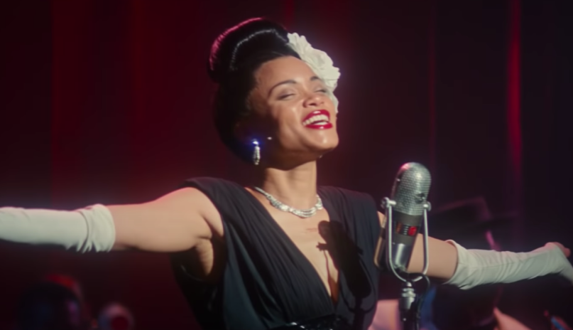 Image of Lady day performing, singing with her eyes closed and hands in the air. She has a red dress and a flower in her hair.