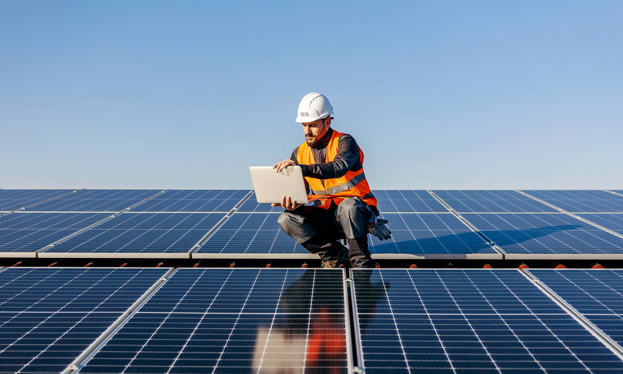 Main in a hard hat sat on solar panels with a laptop.