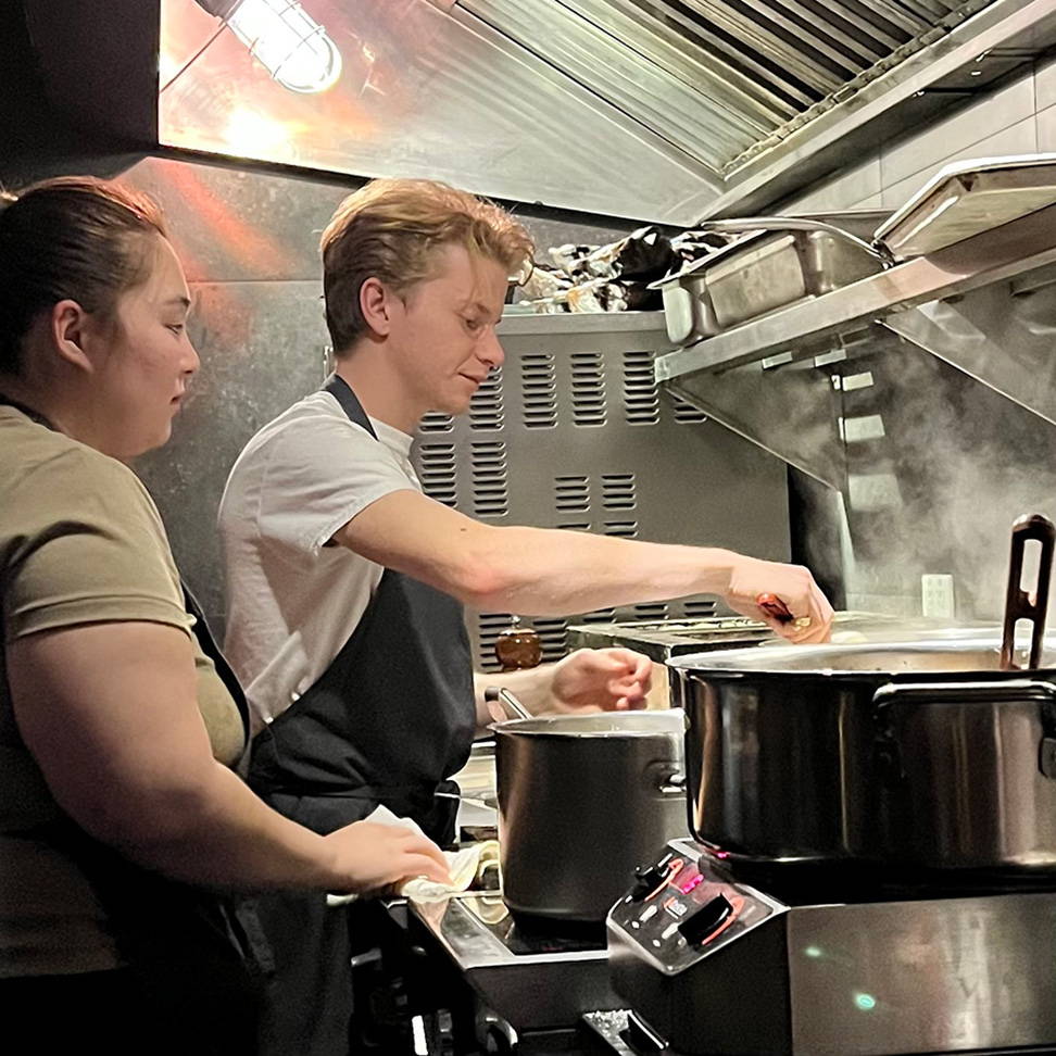 A man and a woman cooking at a stove in a commercial kitchen, the man is stirring a pot, and two steaming pots are in the foreground.