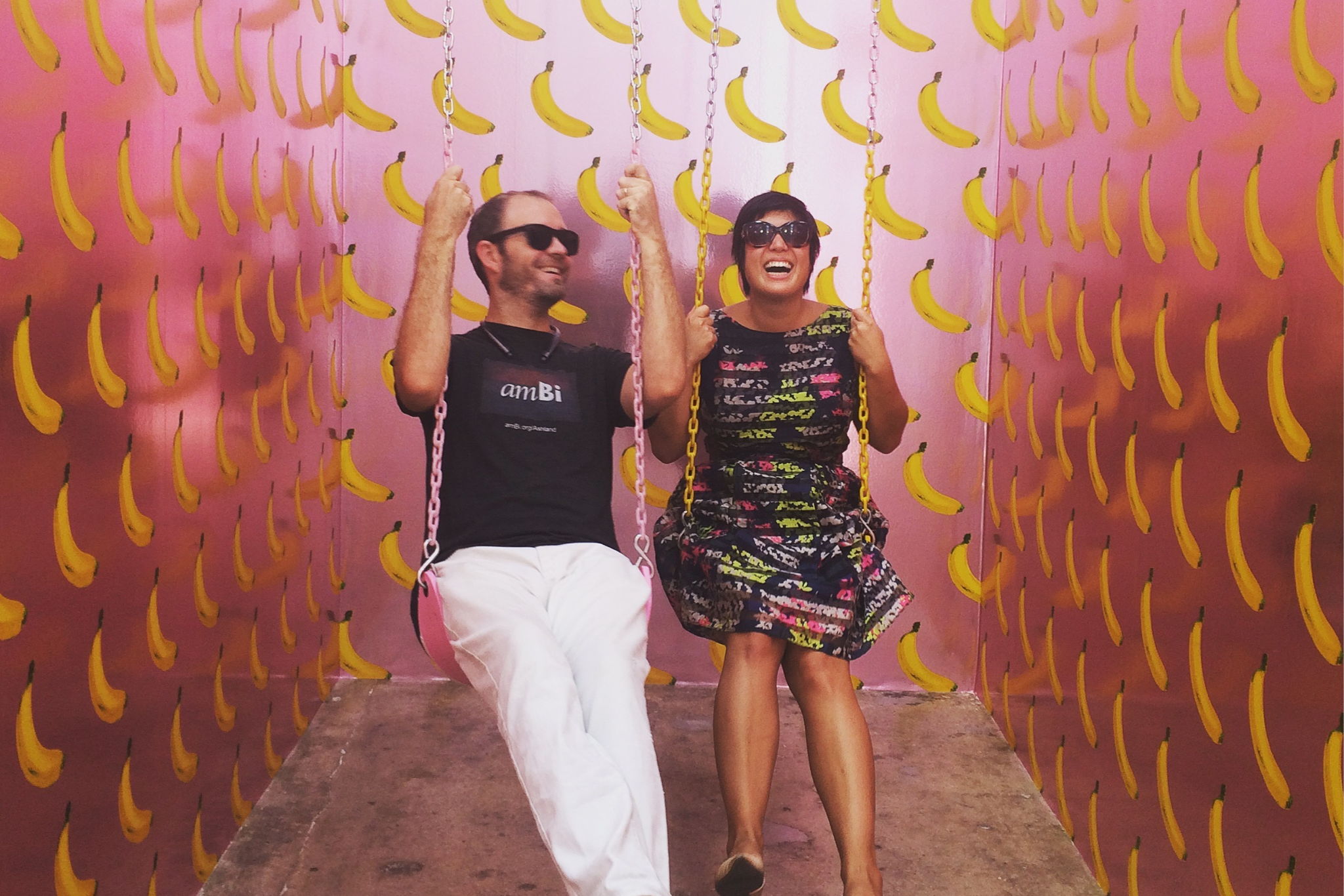 Talia and Rio laughing while on a set of swings inside a pink room with cartoon banana drawing wallpaper.