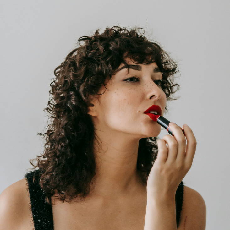 woman with dark curls in a shag haircut, applying lipstick