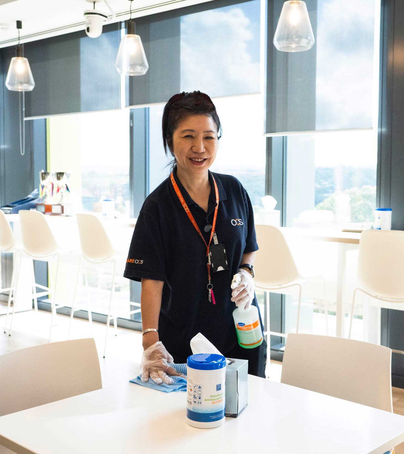 A woman wearing gloves and a dark uniform is smiling while cleaning a table in a modern, bright kitchen area with large windows and white chairs. Cleaning supplies are on the table.