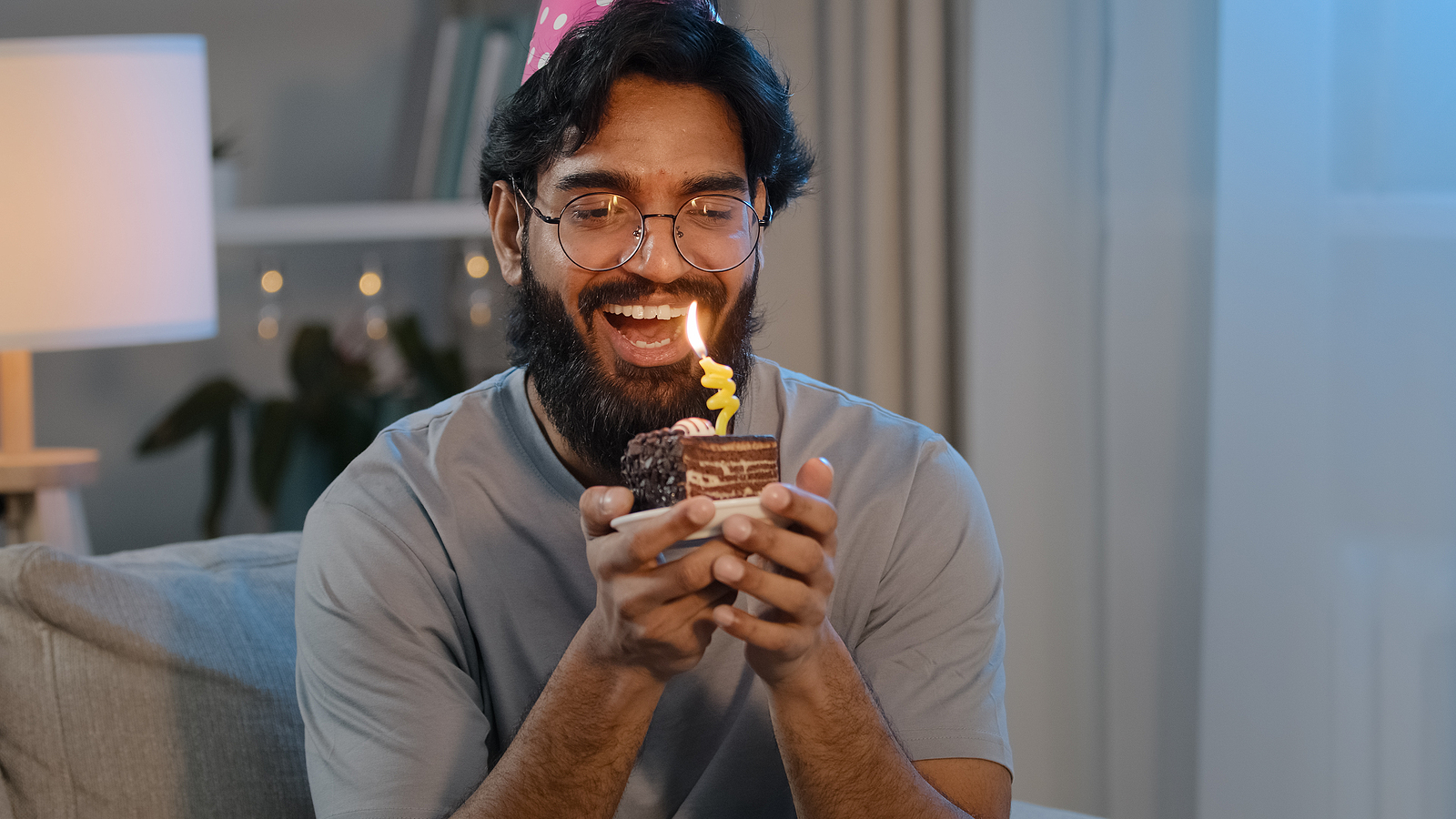 happy indian male face bearded man wears festive birthday pink hat and glasses holds cupcake makes wish joyful congratulating at home blowing burning candles