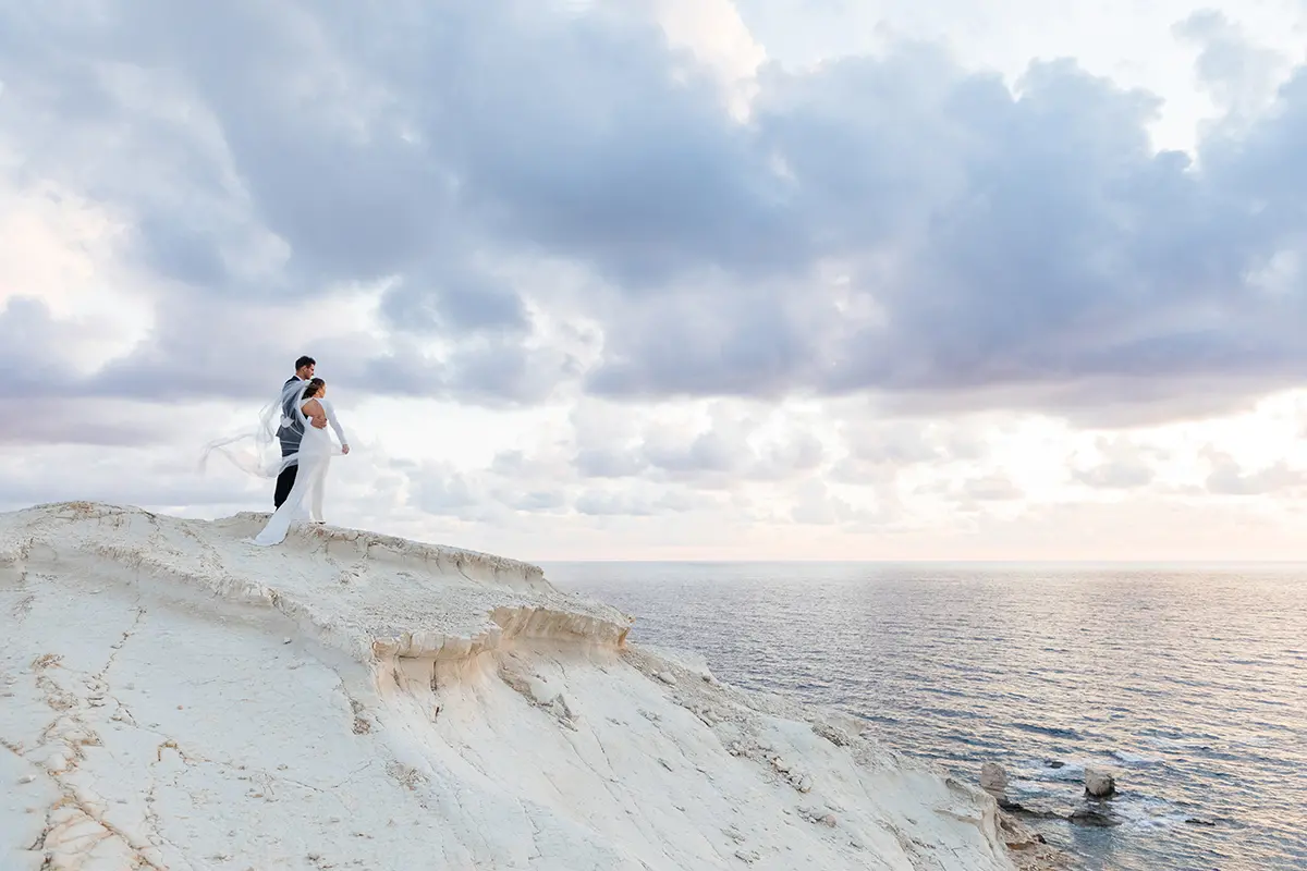 Un photographe de mariage capture le premier regard du couple sur une falaise isolée surplombant les eaux bleues claires à Paphos.