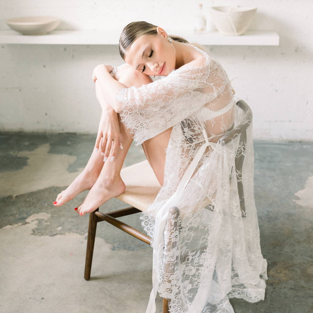 a model poses for a bridal editorial on a chair wearing By Catalfo's long,  lace Lily Robe
