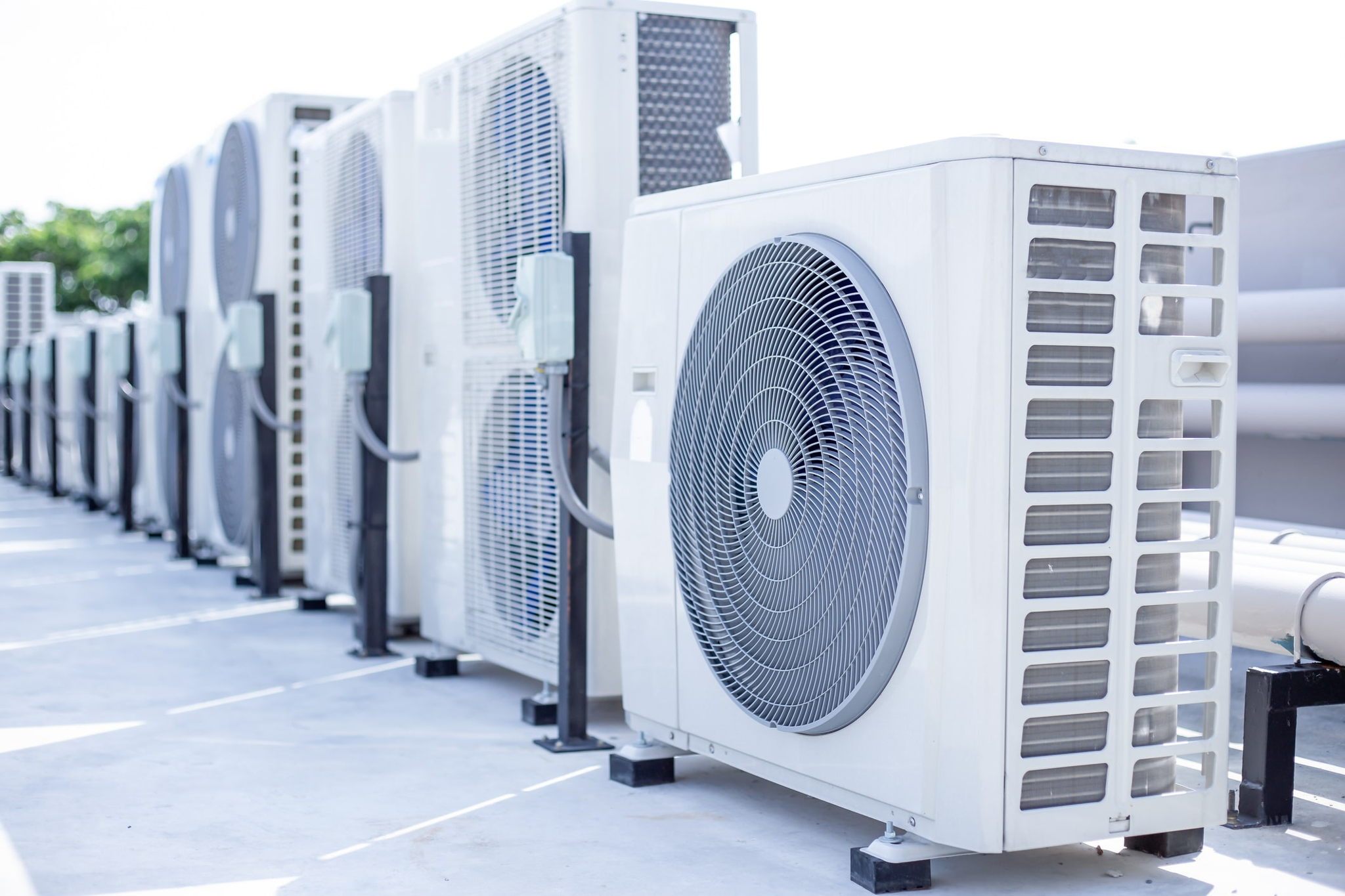 A row of outdoor air conditioning units lined up on a rooftop, with white metal casings and large circular fans visible on the front panels. The units are installed on raised black platforms.