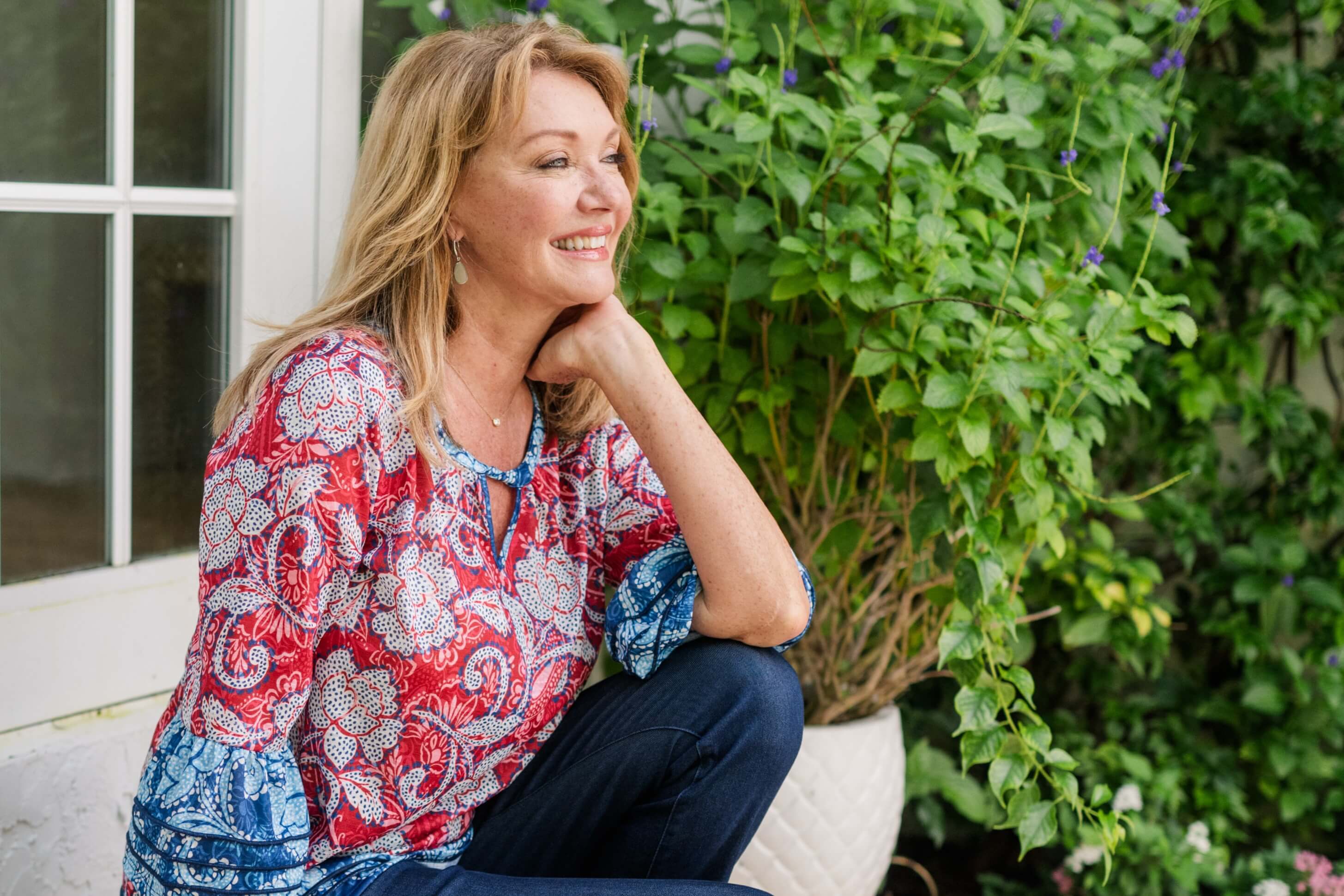 A woman in a printed blouse and dark jeans sits on the stairs of a stoop in a garden
