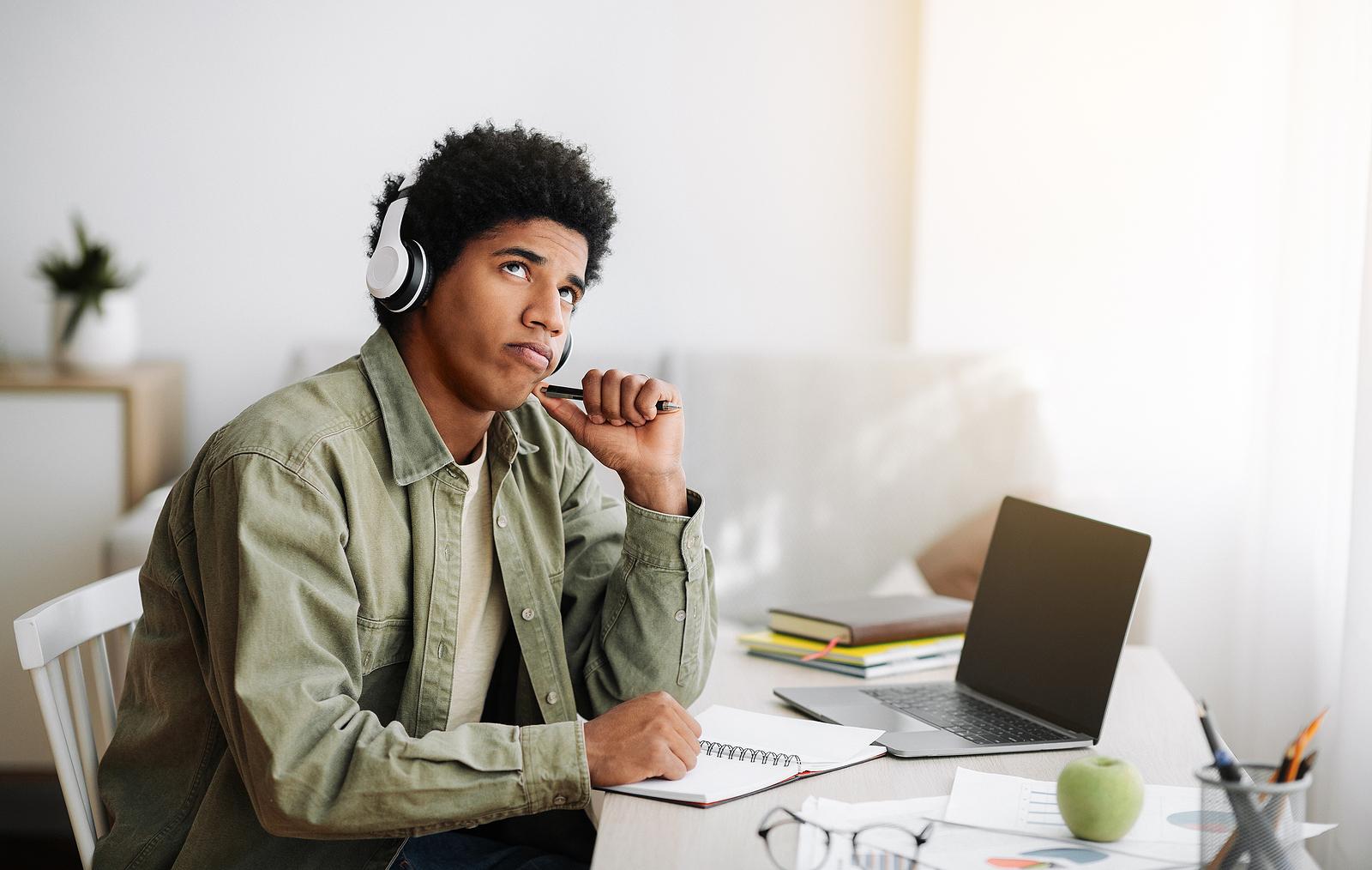 A young black man is deep in thought looking at the ceiling while working on his computer and notepad.