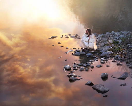 Jesus crouched on a rocky shore looking out at a lake reflecting the orange and pink clouds.