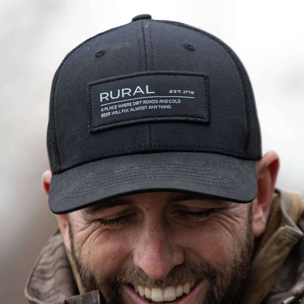 A smiling man wearing a black hat with a patch that reads, Rural: A place where dirt roads and cold beer will fix almost anything. This represents the Rural Definition Collection