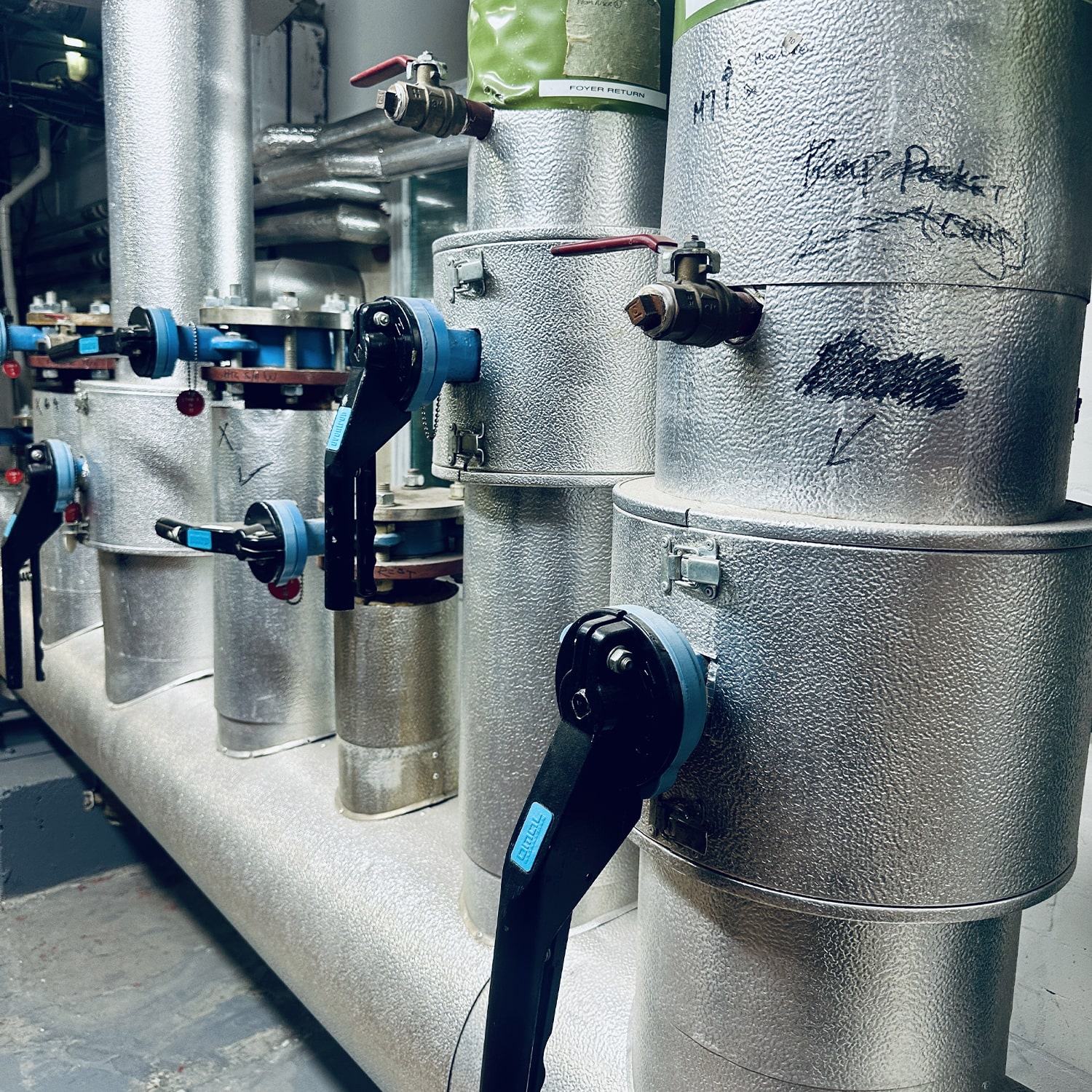 Close-up of insulated metal pipes and valves in an industrial setting, with blue-handled levers and handwritten notes and markings on the surface.