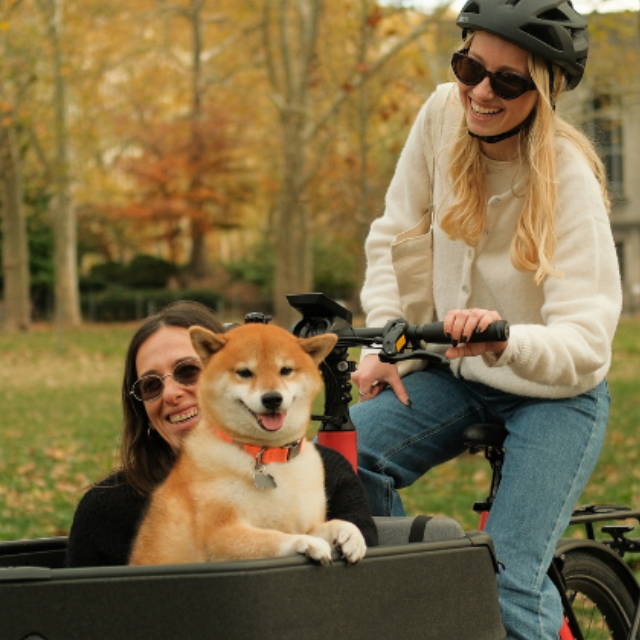 two women and a Shiba Inu dog riding a front loader electric cargo bike in the park