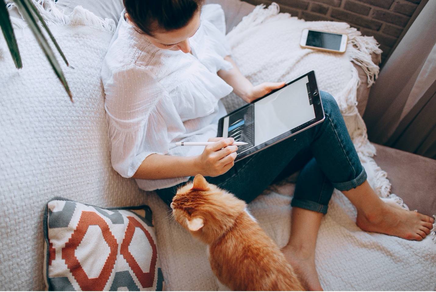 Women writing on her tablet while sitting on the couch next to her orange cat