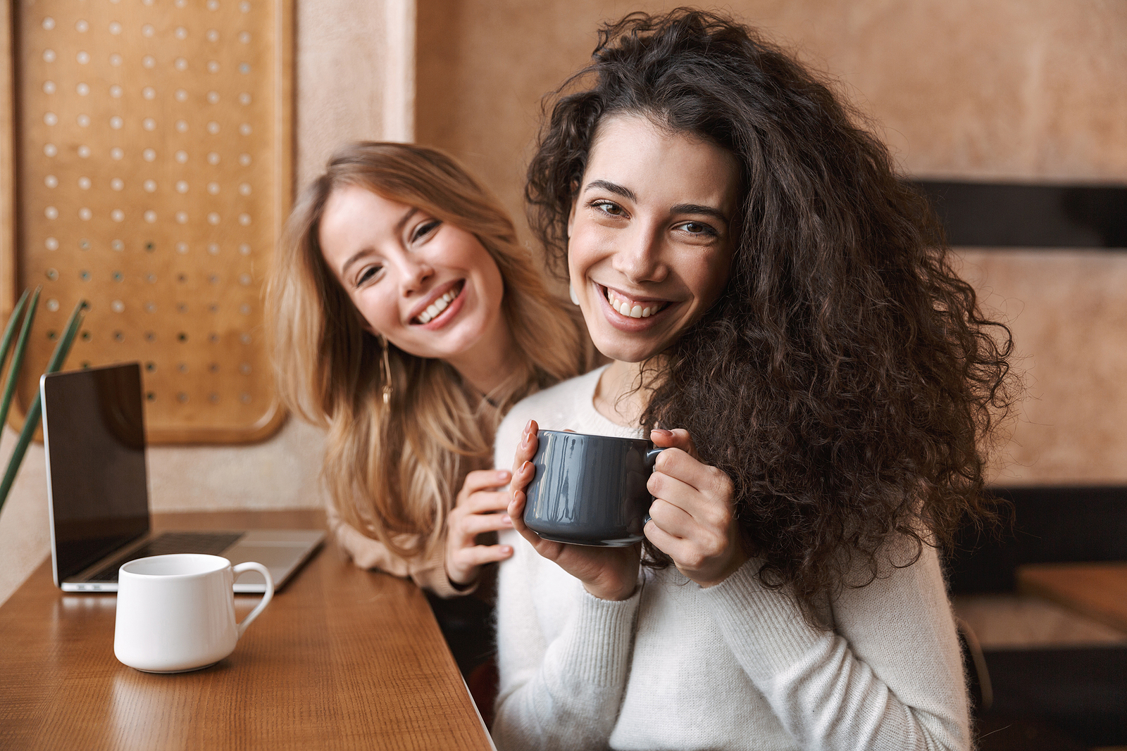 Two cheerful young girlfriends sitting at the cafe indoors, having cup of coffee.