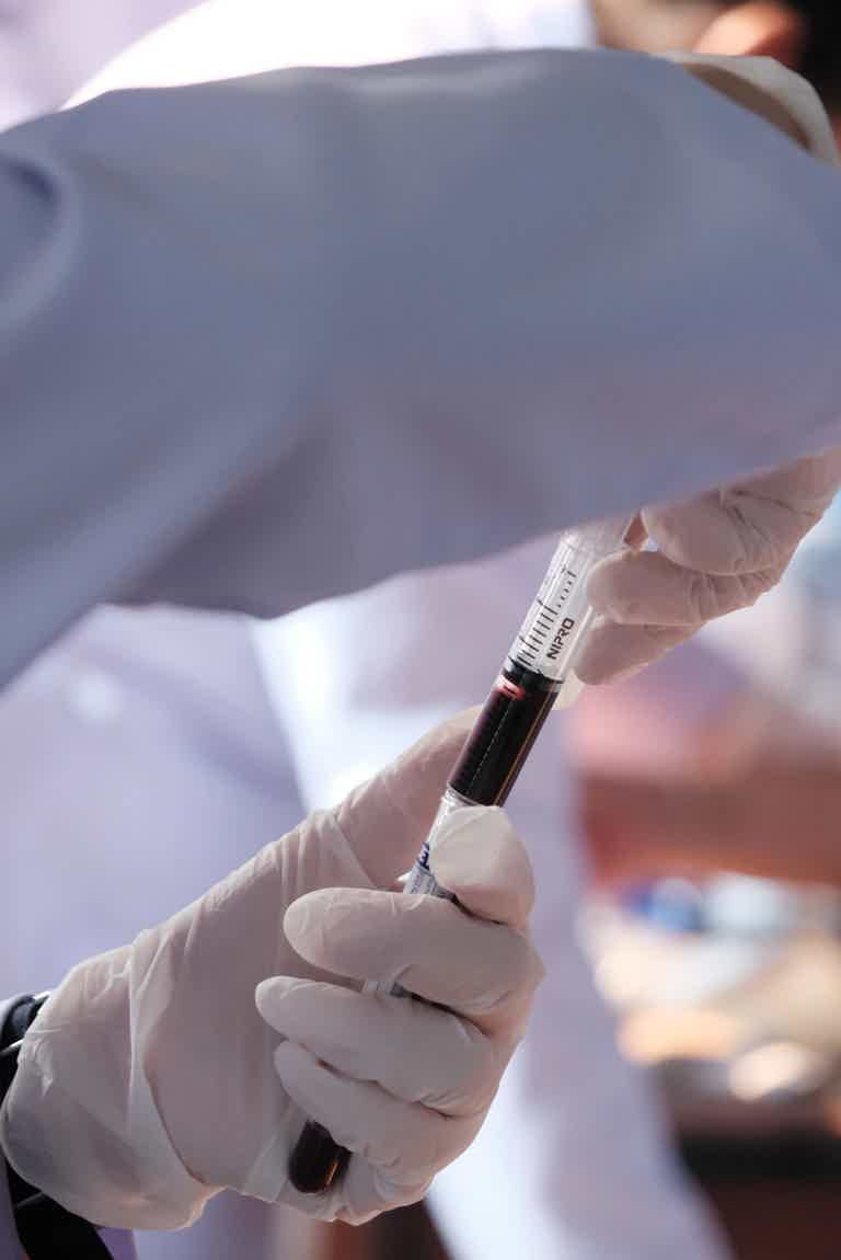 Close-up of gloved hands holding a syringe and vial, drawing dark red blood in a medical or laboratory setting.