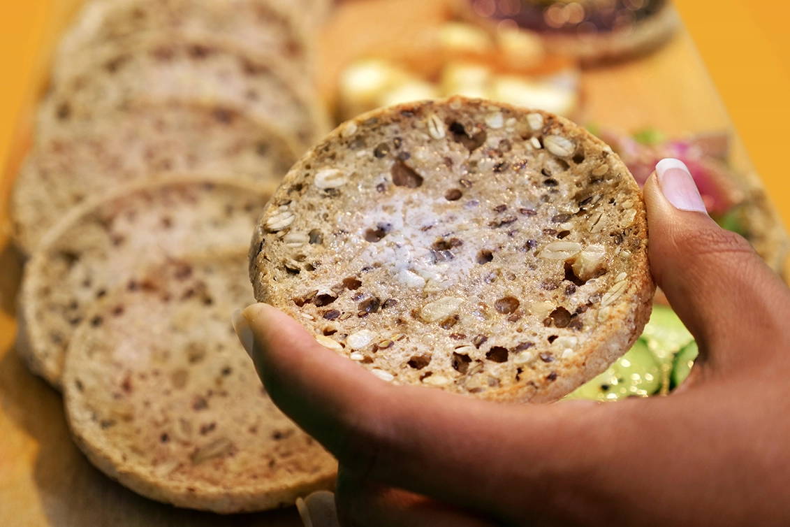 A close-up of a hand holding a toasted multi-seed English muffin with melted butter, with a row of sliced English muffins blurred in the background.