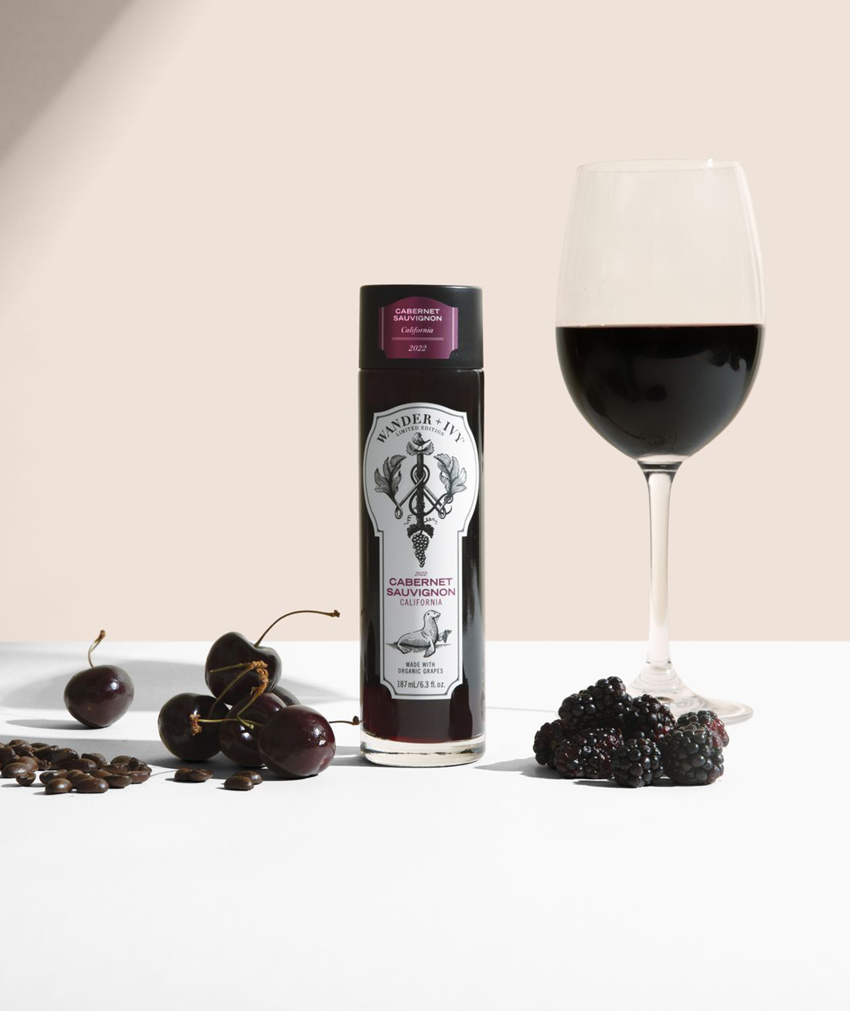 A single-serve bottle of Cabernet Sauvignon next to a filled wine glass, cherries, coffee beans, and blackberries