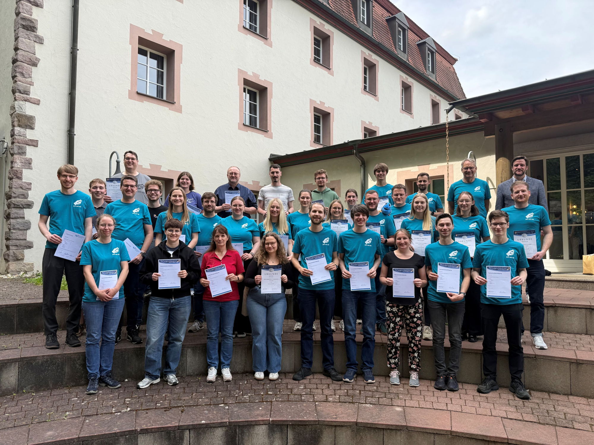 Auf einer Treppe stehen viele Menschen und halten Urkunden vor sich. Einige von ihnen tragen einheitliche türkise T-Shirts. 