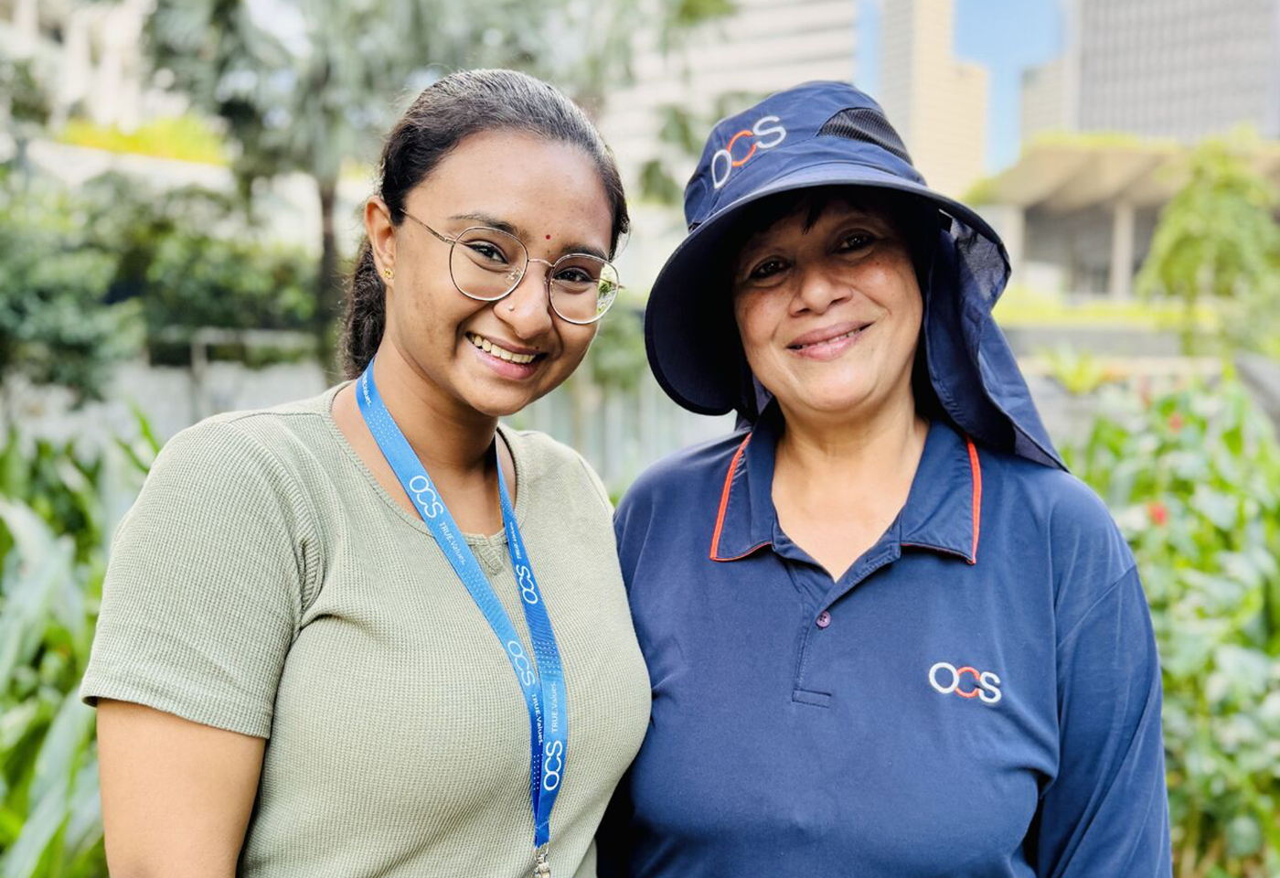 Two women smiling outdoors, one wearing glasses and an OCS lanyard, the other in an OCS uniform and hat. Greenery and tall buildings are visible in the background.